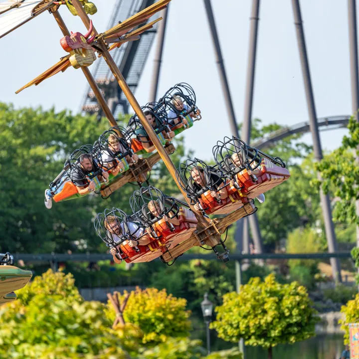 Besucher schweben mit der Attraktion "Hicks Himmelstürmer" im Heide Park Resort liegend durch die Lüfte. Im Hintergrund sind Bäume zu sehen und die Attraktion KRAKE.
